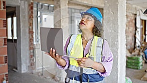 Young beautiful latin woman builder using laptop looking around at construction site