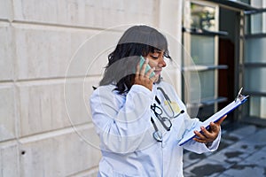 Young beautiful latin woman doctor talking on smartphone reading document at hospital