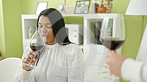 Young beautiful latin woman drinking glass of wine sitting on table at home