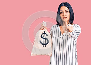 Young beautiful latin woman holding money bag with dollar symbol pointing with finger to the camera and to you, confident gesture