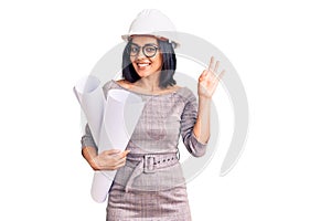 Young beautiful latin woman wearing architect hardhat and glasses holding blueprints doing ok sign with fingers, smiling friendly