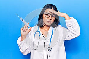 Young beautiful latin woman wearing doctor stethoscope holding syringe stressed and frustrated with hand on head, surprised and