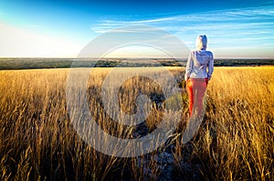 Young beautiful lean girl wears red pants and white hoodie stays in a field among golden grass at sunset, back to camera