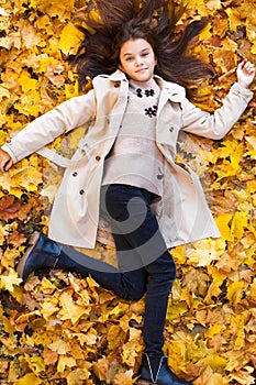 Young beautiful little girl in beige coat lying on yellow leaves