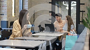 Young beautiful lonely woman drinking coffee in cafe with blurred happy couple talking and hugging at the background