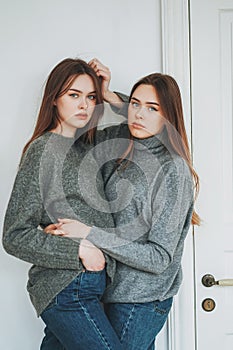 Young beautiful long hair girls sisters twins in casual grey sweaters and jeans on background of the white door