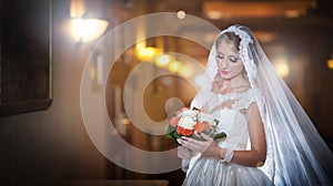 Young beautiful luxurious woman in wedding dress posing in luxurious interior. Bride with long veil holding her wedding bouquet