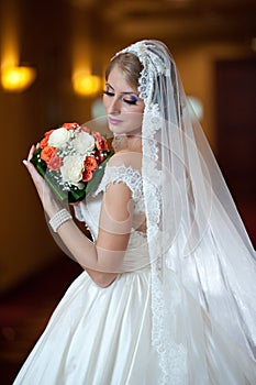 Young beautiful luxurious woman in wedding dress posing in luxurious interior. Bride with long veil holding her wedding bouquet