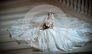 Young beautiful luxurious woman in wedding dress sitting on stair steps in semi-darkness. Bride with huge wedding dress
