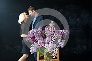 Young beautiful man in dark blue suit and woman in black dress embracing each other and lilac bouquet in fokus