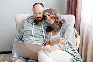 Young beautiful married couple are sitting on armchair at home interior using laptop. Happy smiling Woman and man do