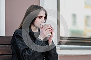 Young beautiful millenial girl in a coat is sitting on an outdor bench with a paper cup of coffee. Autumn or spring day