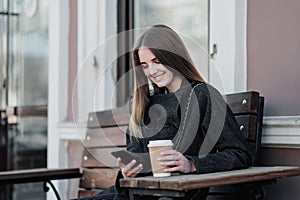 Young beautiful millenial girl in a coat is sitting on an outdor bench with a paper cup of coffee. Autumn or spring day