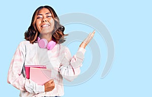 Young beautiful mixed race woman holding books wearing headphones celebrating victory with happy smile and winner expression with