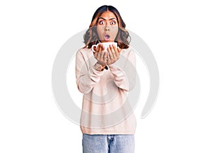 Young beautiful mixed race woman holding a cup of coffee scared and amazed with open mouth for surprise, disbelief face