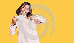 Young beautiful mixed race woman holding a cup of coffee smiling happy pointing with hand and finger