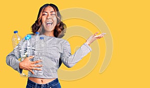 Young beautiful mixed race woman holding recycling plastic bottles celebrating victory with happy smile and winner expression with