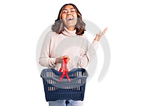 Young beautiful mixed race woman holding supermarket shopping basket celebrating victory with happy smile and winner expression