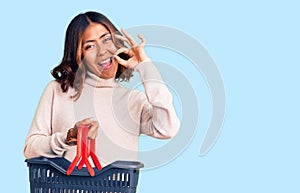 Young beautiful mixed race woman holding supermarket shopping basket doing ok sign with fingers, smiling friendly gesturing