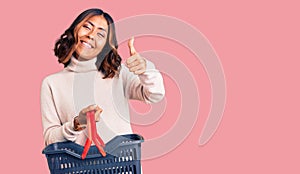 Young beautiful mixed race woman holding supermarket shopping basket smiling happy and positive, thumb up doing excellent and