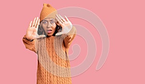 Young beautiful mixed race woman wearing wool sweater and winter hat doing frame using hands palms and fingers, camera perspective