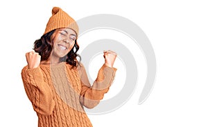 Young beautiful mixed race woman wearing wool sweater and winter hat very happy and excited doing winner gesture with arms raised,