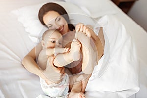 Young beautiful mom and newborn baby lying in bed smiling fooling at home. From above.