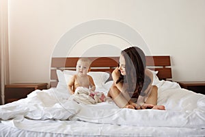 Young beautiful mom in sleepwear sitting on bed with her baby daughter smiling playing at home.