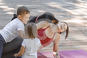 Young beautiful mother with baby son, daughter and with dog exercising and doing yoga at summer park at sunrise