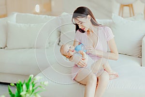 Young beautiful mother bottle feeding her little son at home