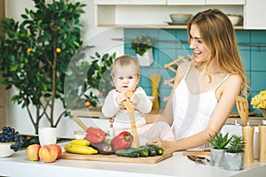 Young beautiful mother is cooking and playing with her baby daughter in a kitchen setting. Healthy food concept