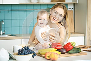 Young beautiful mother is cooking and playing with her baby daughter in a modern kitchen setting. Healthy food concept. Taking