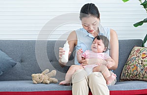 Young beautiful mother holding and feeding milk to cute little Caucasian 7 months newborn baby, sitting together on sofa at home