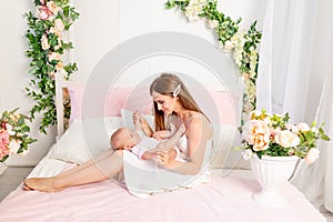 A young beautiful mother holds her daughter a girl of 6 months on her lap on a white bed, playing and kissing her, place for text