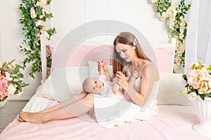 A young beautiful mother holds her daughter a girl of 6 months on her lap on a white bed, playing and kissing her, place for text