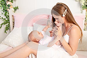 Young beautiful mother holds a little girl daughter 6 months on her lap and kisses her legs on a white bed, mother`s day, place