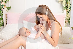 Young beautiful mother holds a little girl daughter 6 months on her lap and kisses her legs on a white bed, mother`s day, place