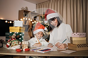 Young beautiful mother and little cute child daughter in red hats writing letter to Santa Claus