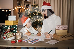 Young beautiful mother and little cute child daughter in red hats writing letter to Santa Claus