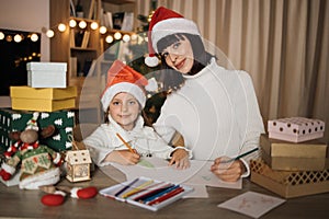 Young beautiful mother and little cute child daughter in red hats writing letter to Santa Claus