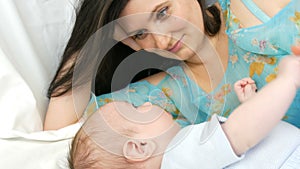Young beautiful mother with long dark hair is lying on a white bed and playing with newborn baby of two months