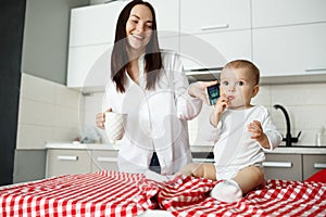 Young beautiful mother smiling, drinking tea and puts phone to baby`s ear to hear father voice. Child sitting on table