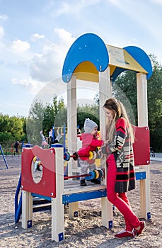 Young beautiful mother in a sweater is playing and riding on a swing with her little baby daughter in a red jacket and hat on the