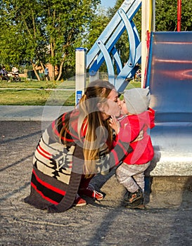 Young beautiful mother in a sweater is playing and riding on a swing with her little baby daughter in a red jacket and hat on the