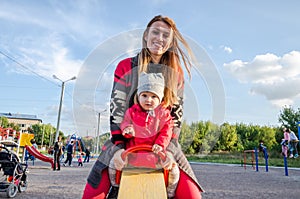 Young beautiful mother in a sweater is playing and riding on a swing with her little baby daughter in a red jacket and hat on the