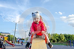 Young beautiful mother in a sweater is playing and riding on a swing with her little baby daughter in a red jacket and hat on the