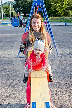 Young beautiful mother in a sweater is playing and riding on a swing with her little baby daughter in a red jacket and hat on the