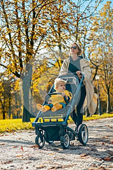 Young beautiful mother wearing a rain coat pushing stroller with her little baby boy child, walking in city park on a