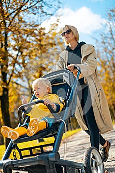 Young beautiful mother wearing a rain coat pushing stroller with her little baby boy child, walking in city park on a