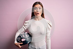 Young beautiful motorcyclist woman with blue eyes holding moto helmet over pink background scared in shock with a surprise face,